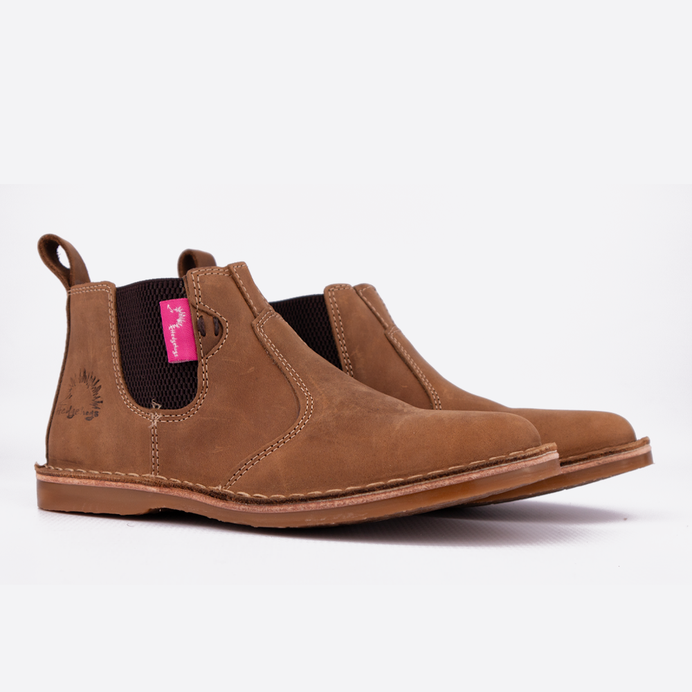 A pair of brown suede Chelsea boots with dark elastic side panels and a bright pink brand tag, shown on a clean white background.