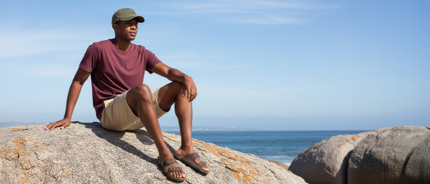 Man wearing green Hippocreek hat sitting on a rock by the ocean with a clear blue sky.