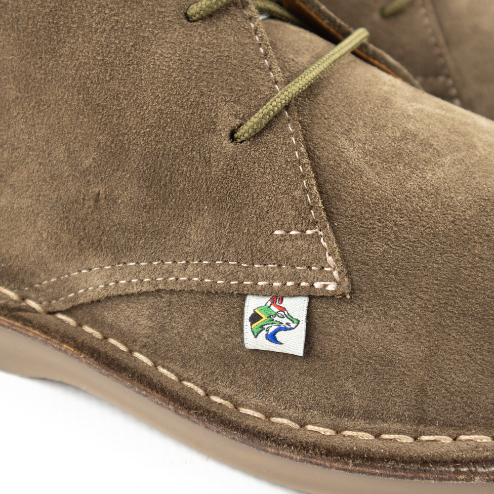 Close-up of an olive suede ankle boot with olive green laces and a small colorful embroidered brand tag near the lace eyelet.