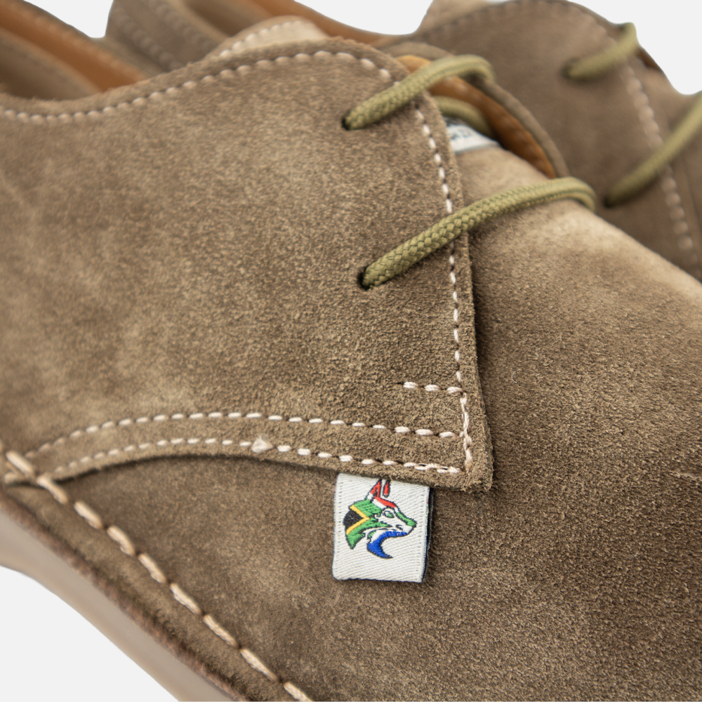 Close-up of an olive-green suede shoe with white stitching, olive green laces, and a small embroidered logo patch on the side.