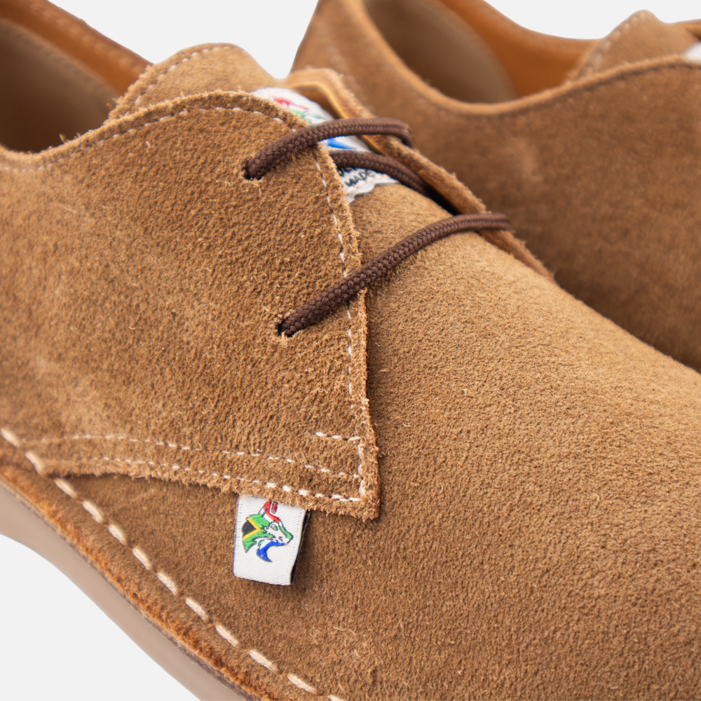 Close-up of a pair of donkey suede lace-up shoes with beige contrast stitching and a small colourful brand tag near the laces.