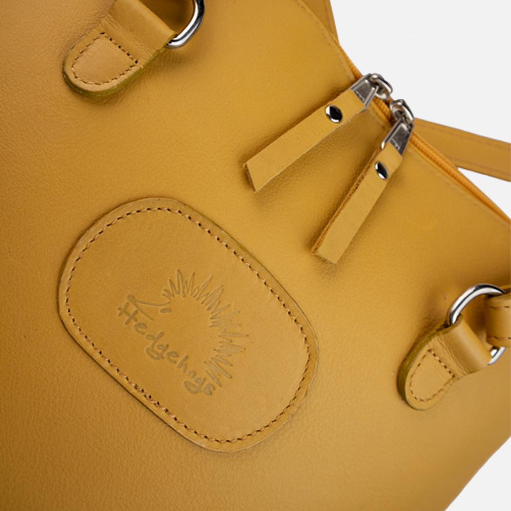 Close up view of the Hedgehogs ladies convertible backpack in ochre yellow leather, showing the embossed logo and double zip detail. 